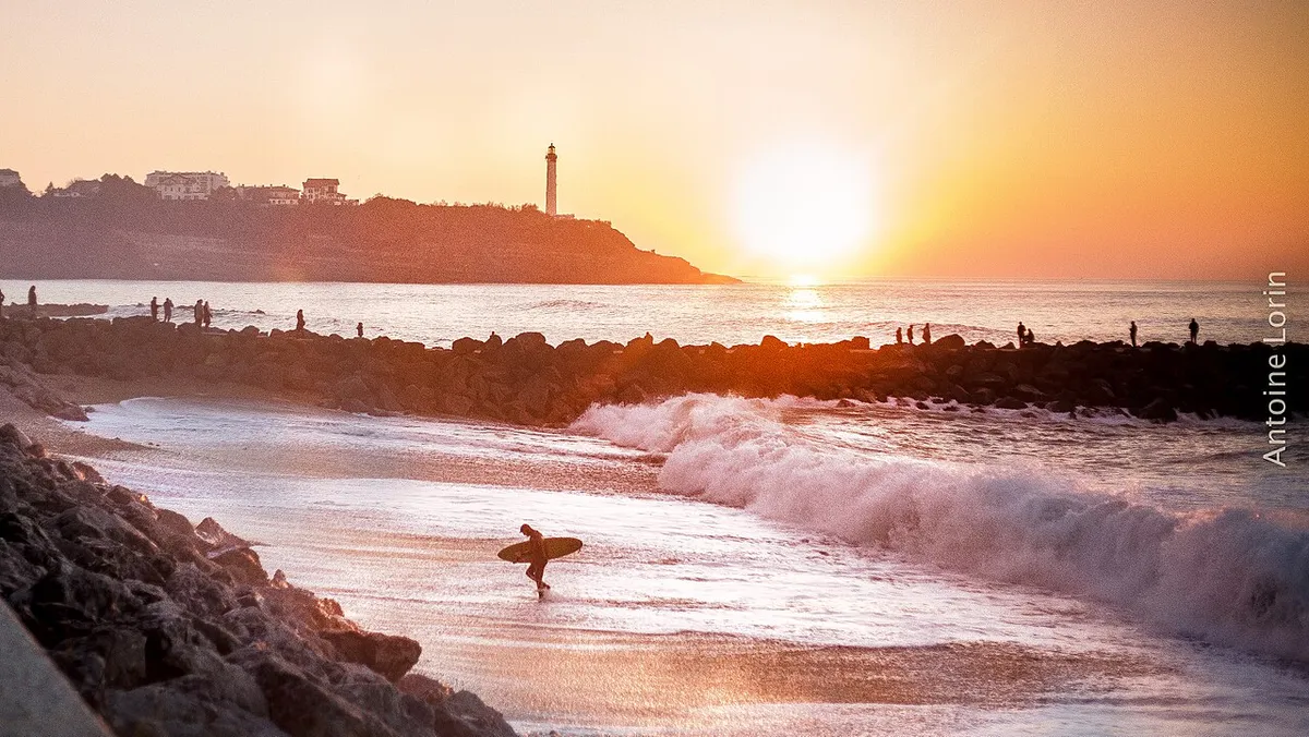 Surfeur sortant de l'eau au coucher du soleil, à côté d'une digue de pierres, avec un phare sur la falaise au loin