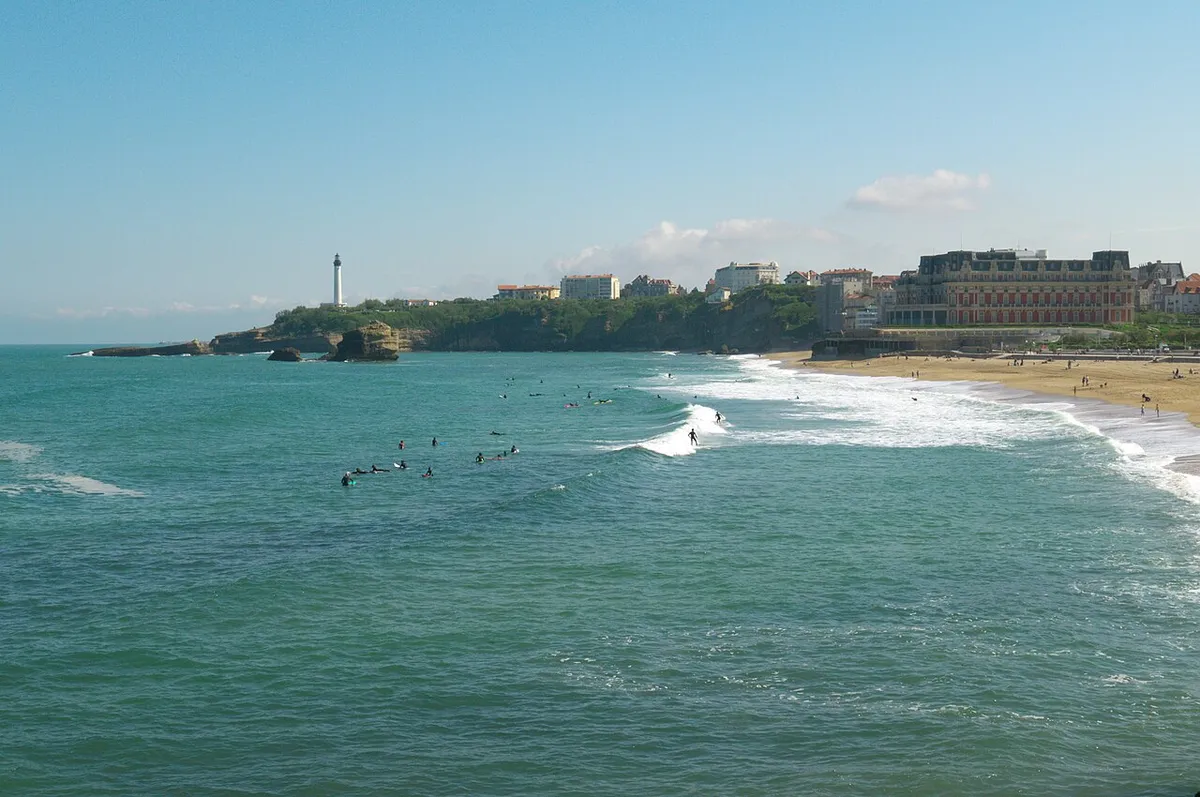 Surfeurs dans l'eau devant la Grande Plage de Biarritz, avec un phare et le front de mer en arrière-plan