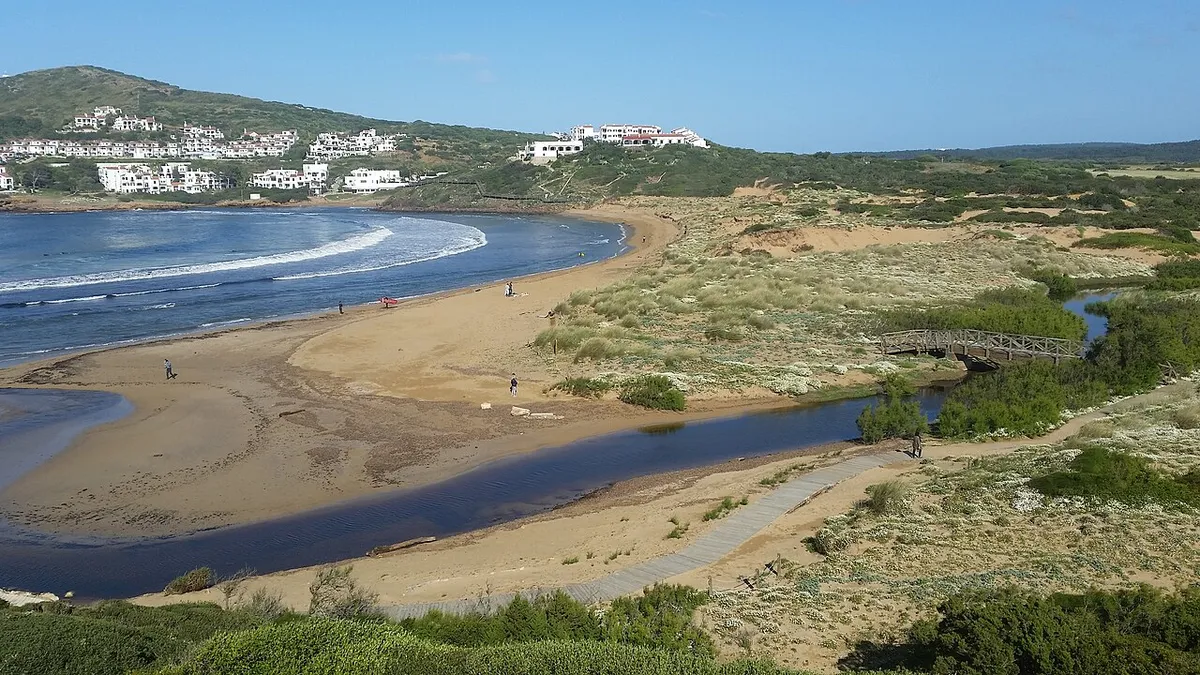 Cala Tirant, à Minorque : une plage en courbe avec des vagues au bord, un ruisseau qui traverse le sable, des dunes au premier plan et des maisons blanches sur la colline derrière