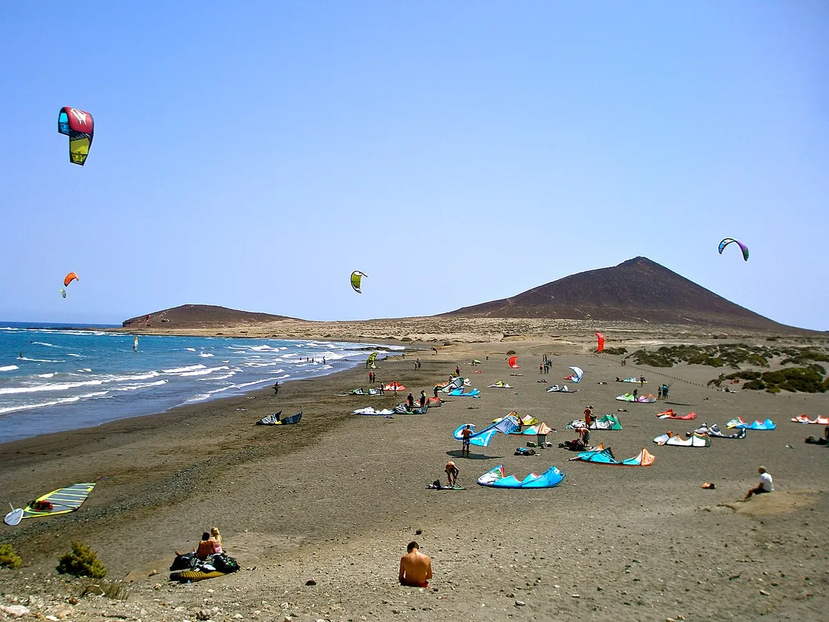 Kites dans le ciel et ailes étalées sur une plage de sable sombre à El Médano, avec une colline conique en arrière-plan