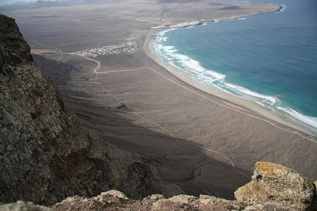Vue plongeante depuis le bord de la falaise sur la longue courbe de la playa de Famara, avec les maisons blanches de l'urbanisation dans les terres