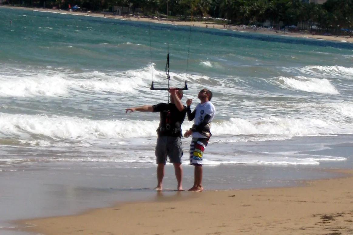 Deux personnes sur la plage tiennent une barre de kite lors d'une leçon, avec des vagues derrière elles
