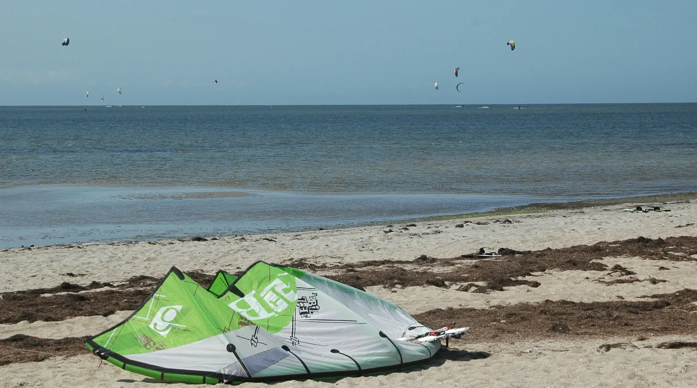 Aile de kitesurf posée sur le sable d'une plage, d'autres ailes en vol au loin