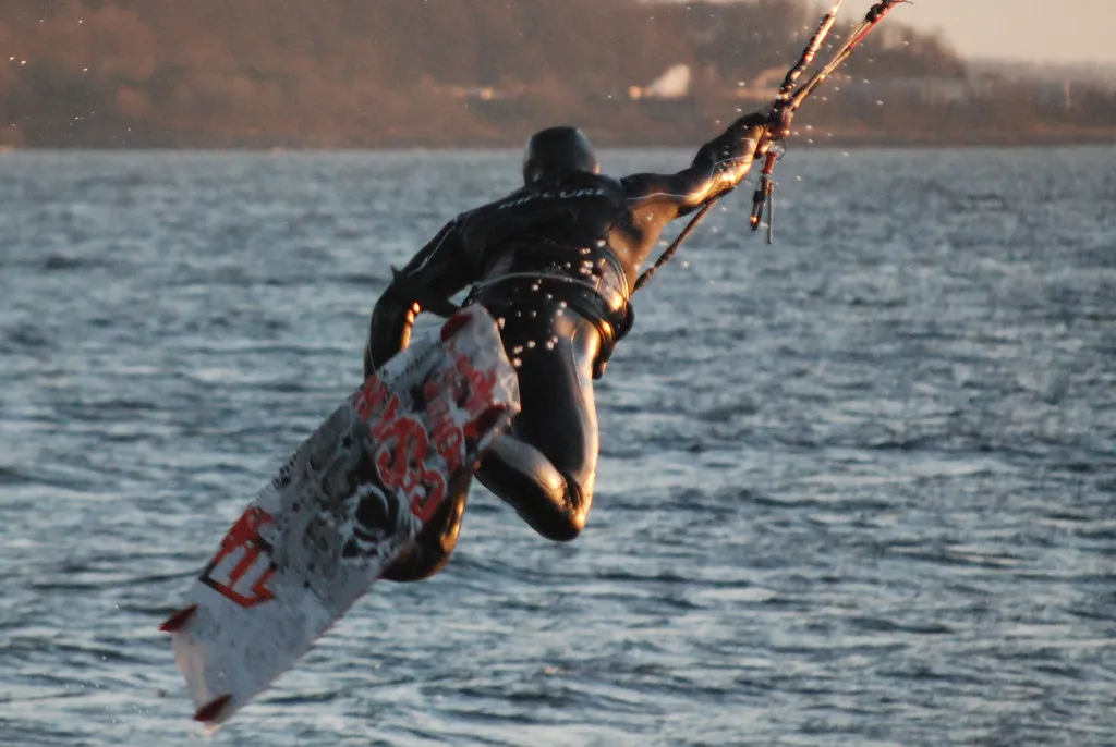 Kitesurfeur en combinaison intégrale noire à capuche, en plein saut, la barre dans une main et la planche sous lui