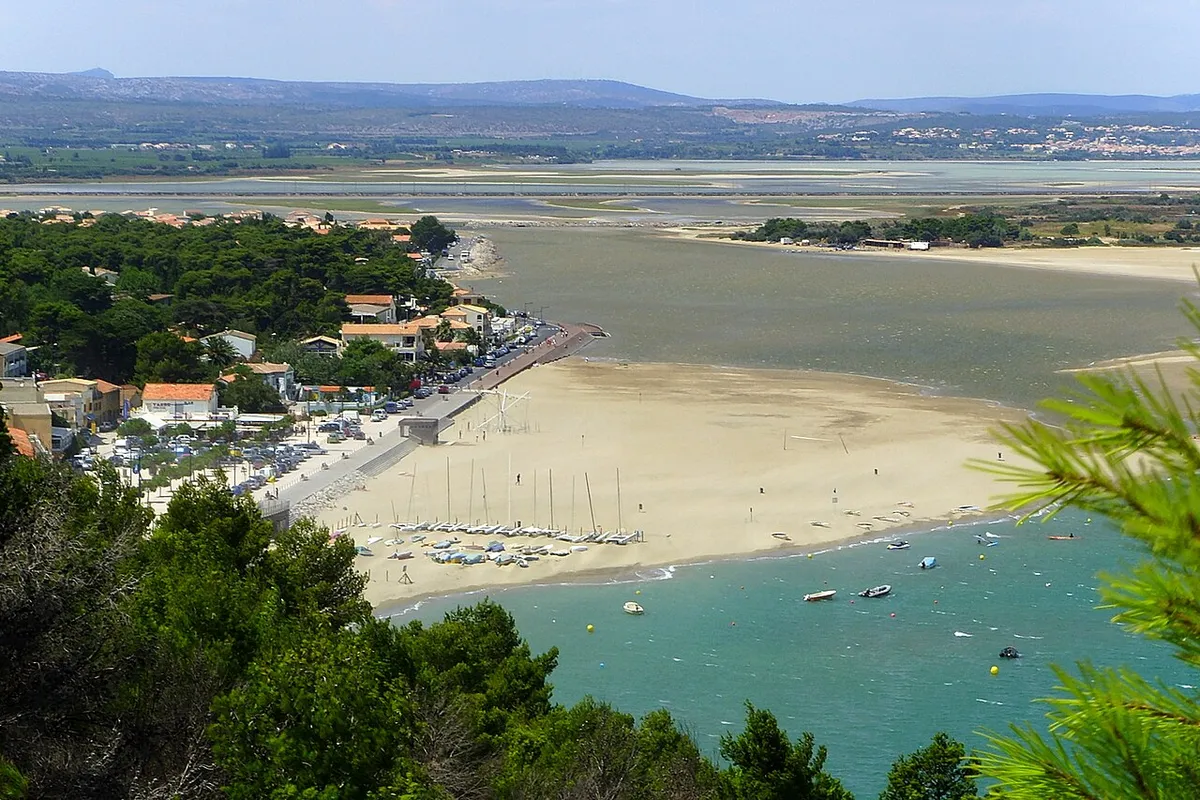 Plage de La Franqui vue depuis la falaise : sable, petits voiliers alignés et eau turquoise, avec l'étang en arrière-plan