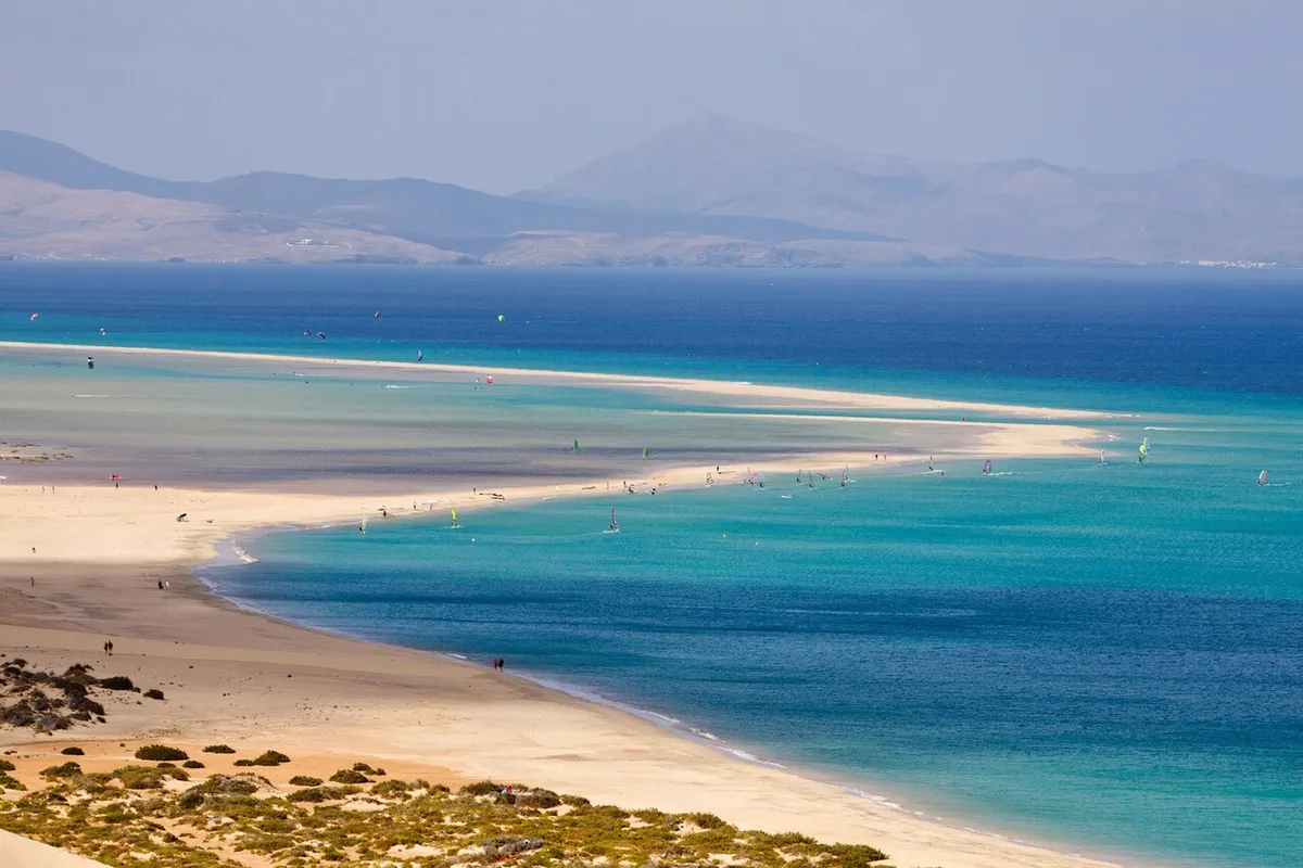 Vue en hauteur du lagon de Sotavento, à Fuerteventura : longue langue de sable, eau turquoise et voiles de windsurf et de kite