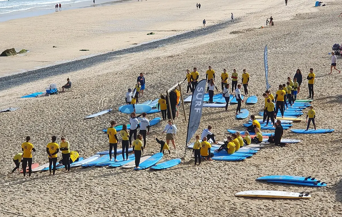 Groupe d'élèves en haut jaune, debout près de planches bleues posées sur le sable pendant un cours de surf collectif