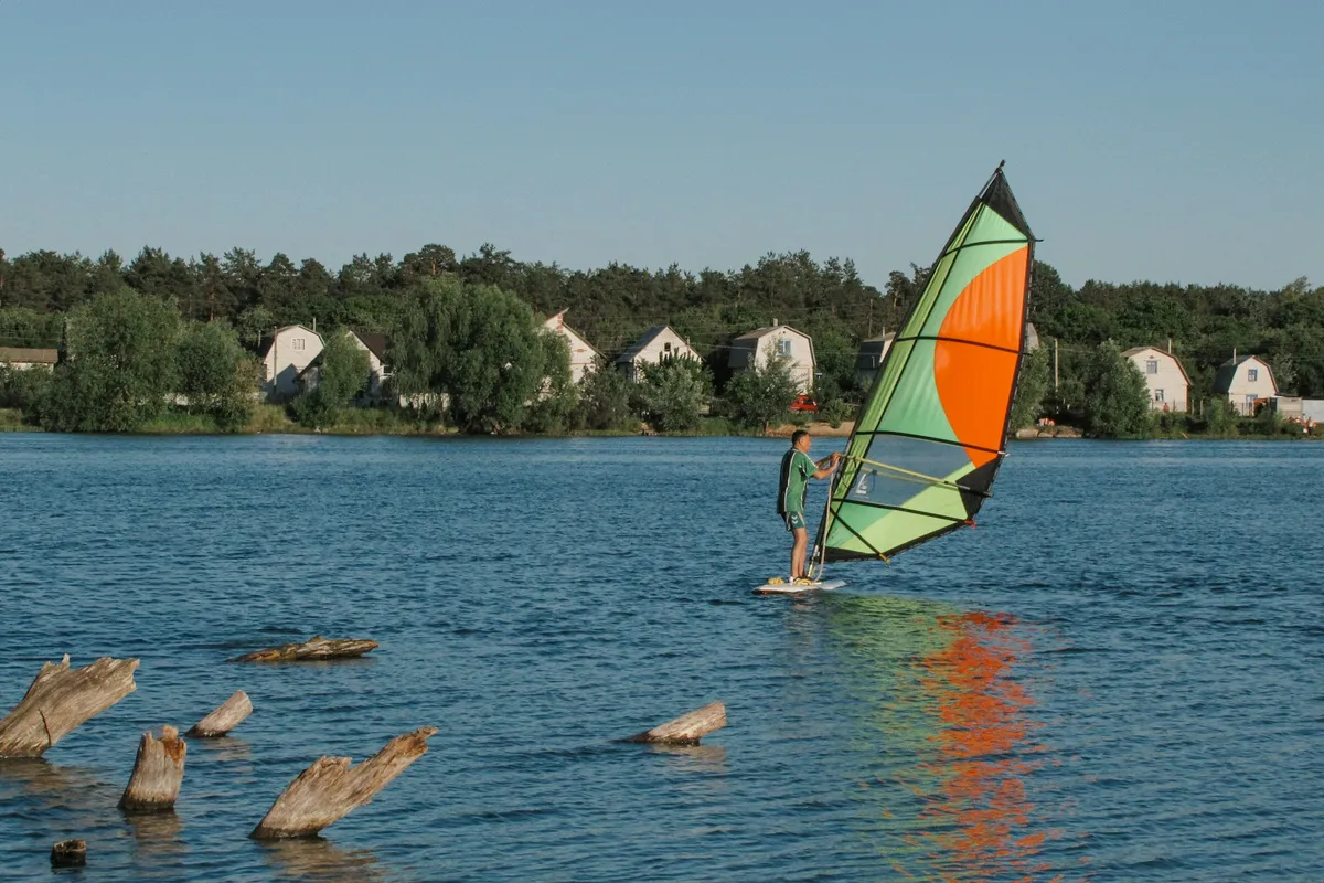 Windsurfeur debout sur sa planche, la voile en main, sur un lac calme bordé d'arbres et de maisons