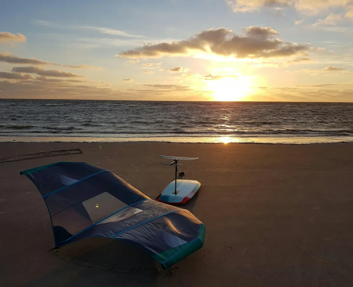 Aile de wing foil posée sur le sable et planche à foil dressée, au coucher du soleil