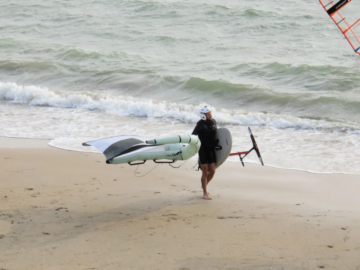 Pratiquant de wing foil casqué portant son aile et sa planche à foil sur la plage