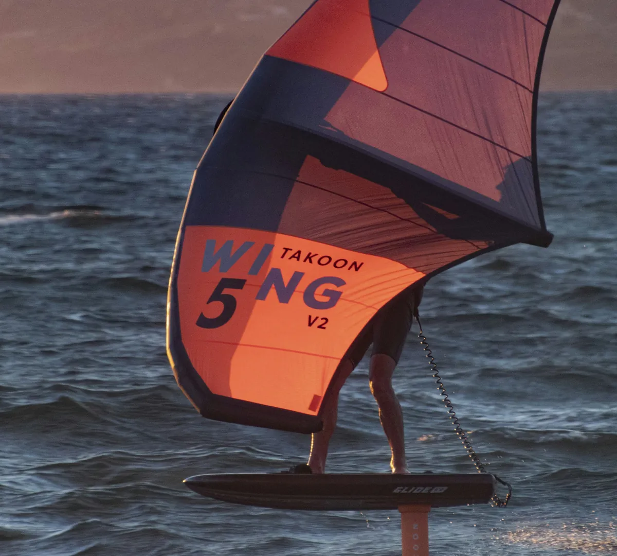 Rider de wing foil derrière une grande aile orange et bleue, planche et mât du foil visibles